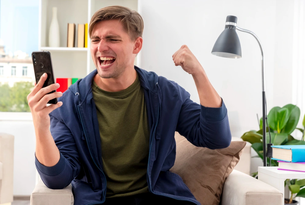 Excited young blonde handsome man sits armchair keeping fist looking phone